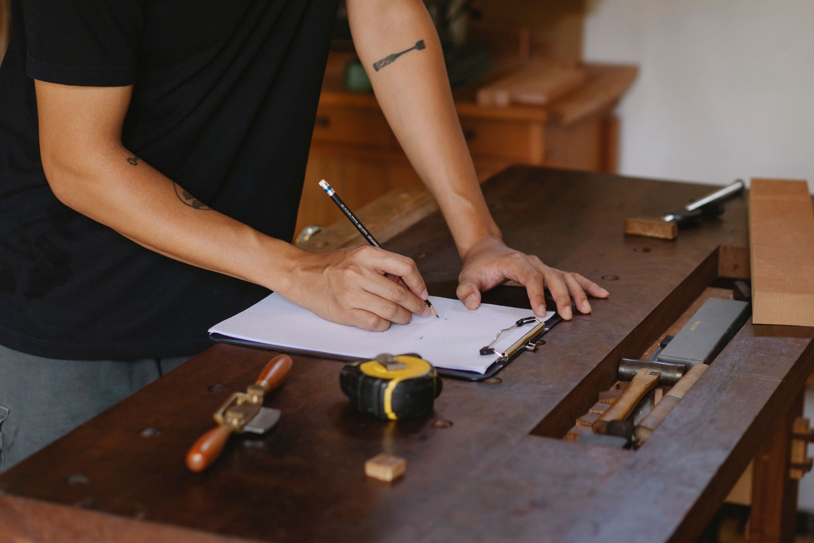 An owner-operator, torso only and no face, writing on a pad at a worn timber bench; a hand plane, a tape measure and chisels laid along the top