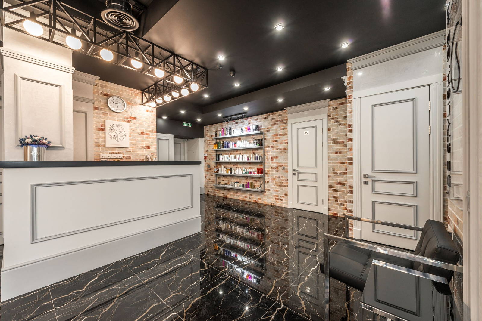 The reception counter of a salon, brick and panelled wall behind, black marble floor reflecting the shelves