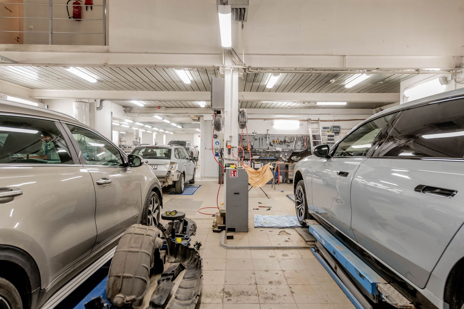 Two cars raised on lifts in a working service bay, a stripped bumper and wheel on the floor between them