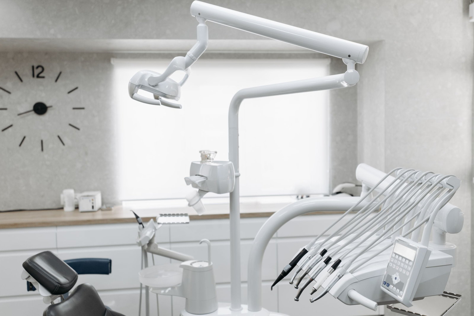 A dental operating light and delivery unit against a bright window, a wall clock on the plaster behind