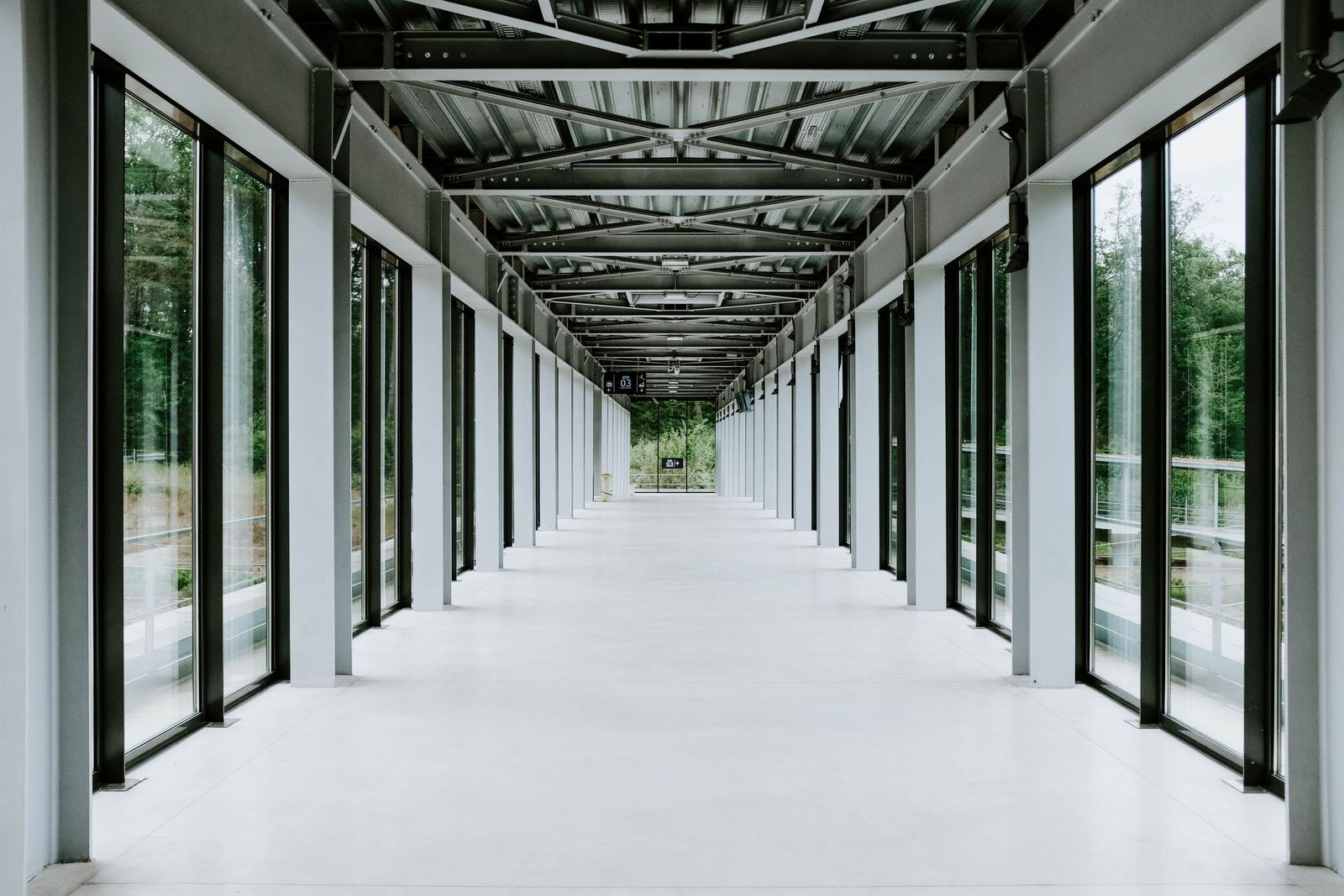 A long glazed walkway, symmetrical steel roof structure, white floor running to a vanishing point, trees outside both sides
