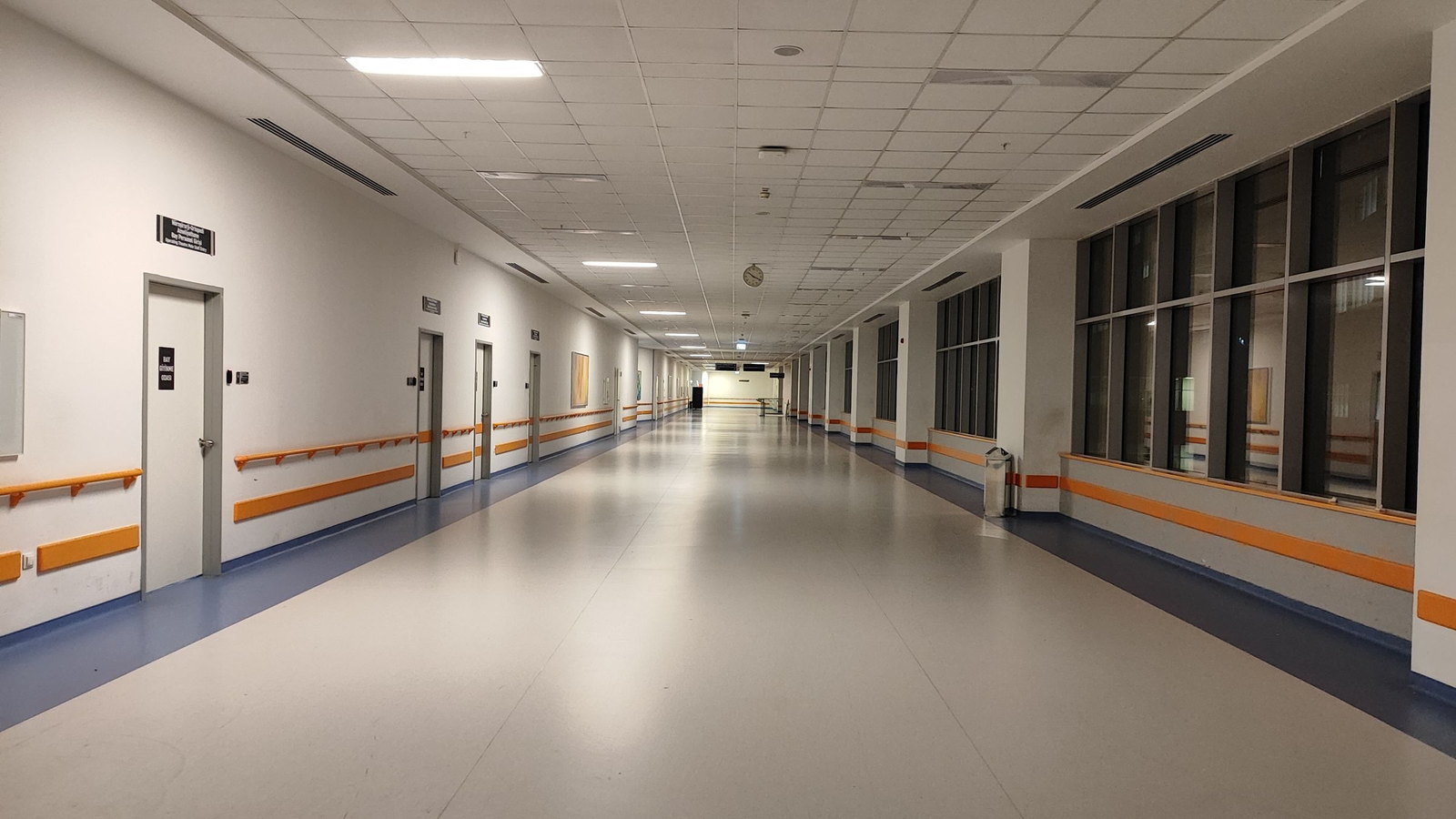 A long, empty hospital corridor with orange handrails, glazing down one side