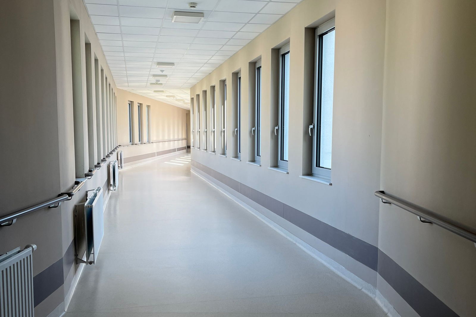 A daylit hospital corridor, windows down one side and handrails running the length of both walls