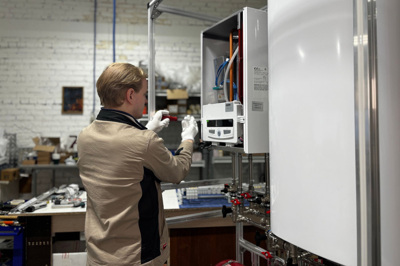 An engineer in workwear, seen from behind, adjusting the valves on an open wall-mounted boiler