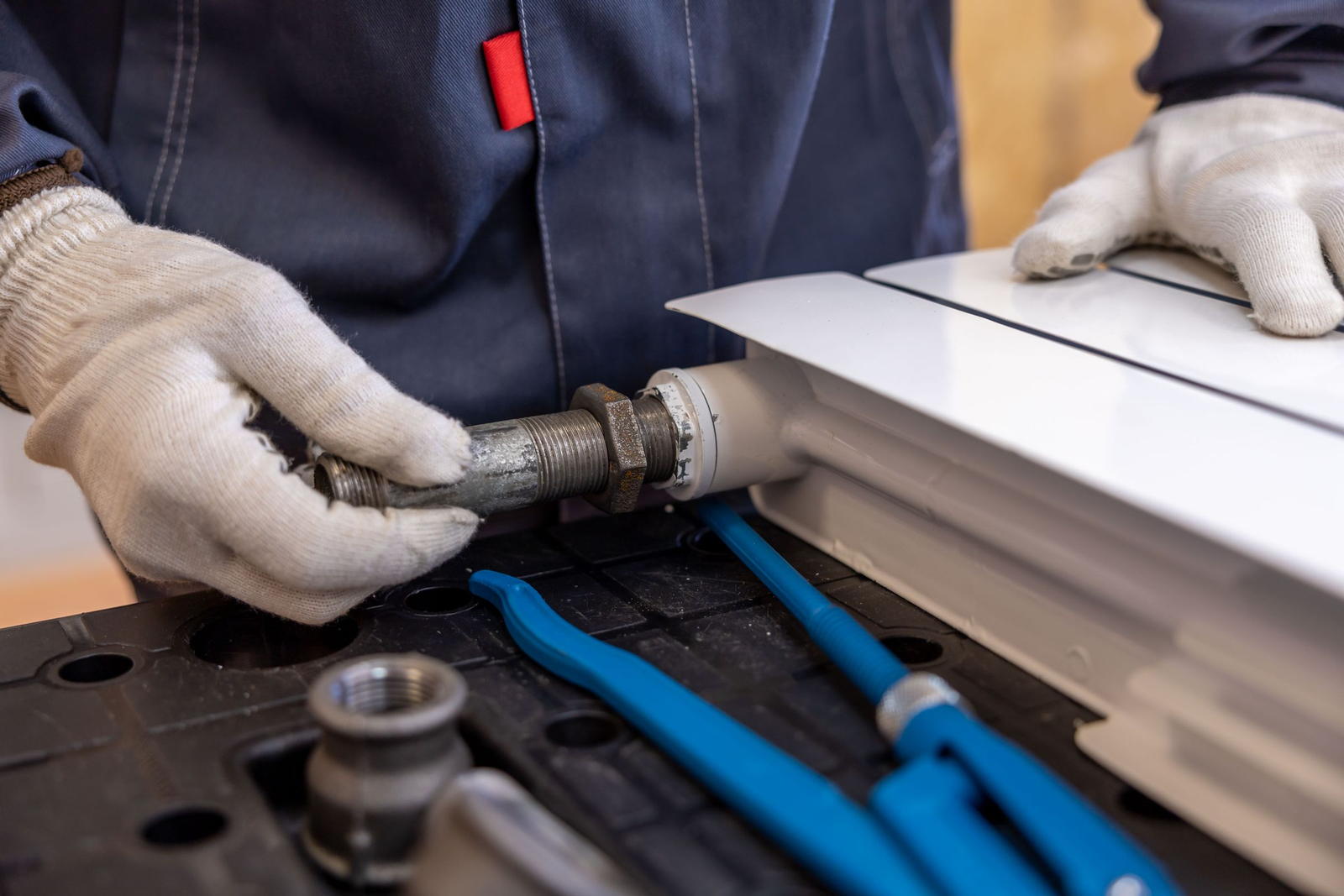 Gloved hands offering a threaded elbow up to a radiator, two pipe wrenches lying on the bench