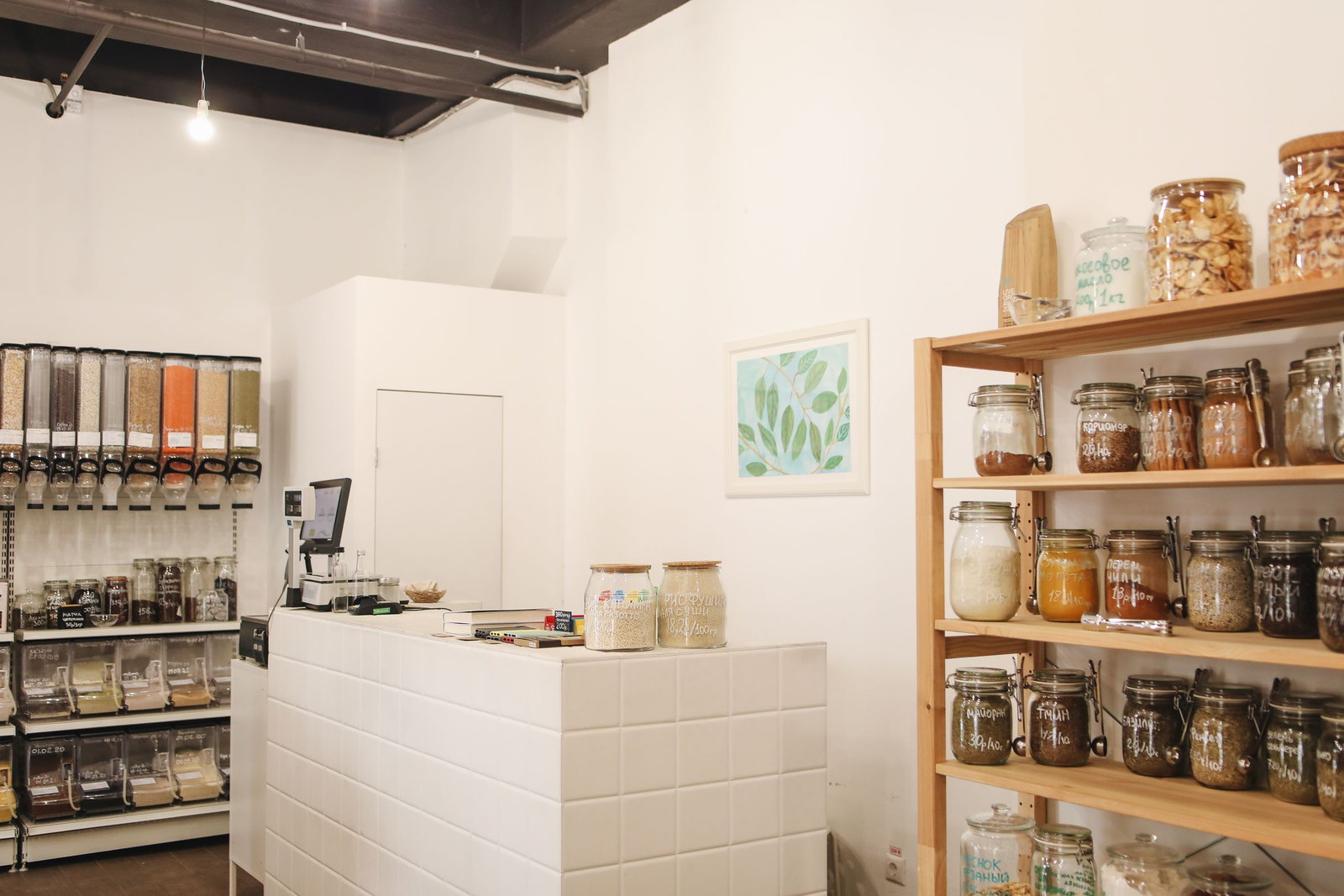 The counter of a refill shop, gravity dispensers on one wall and shelves of labelled jars on the other