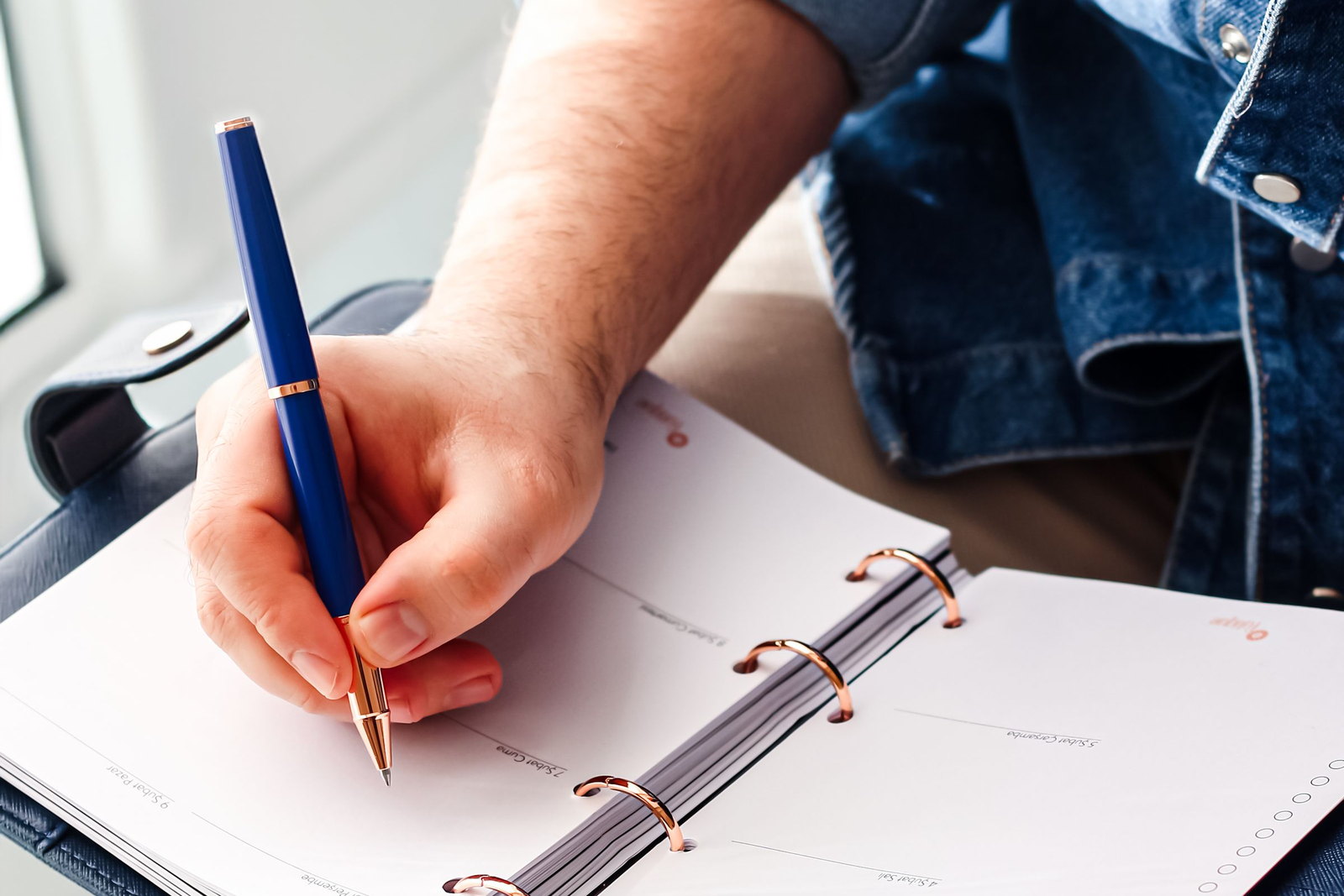 A hand writing into a ring-bound planner balanced on a knee, denim cuff turned back