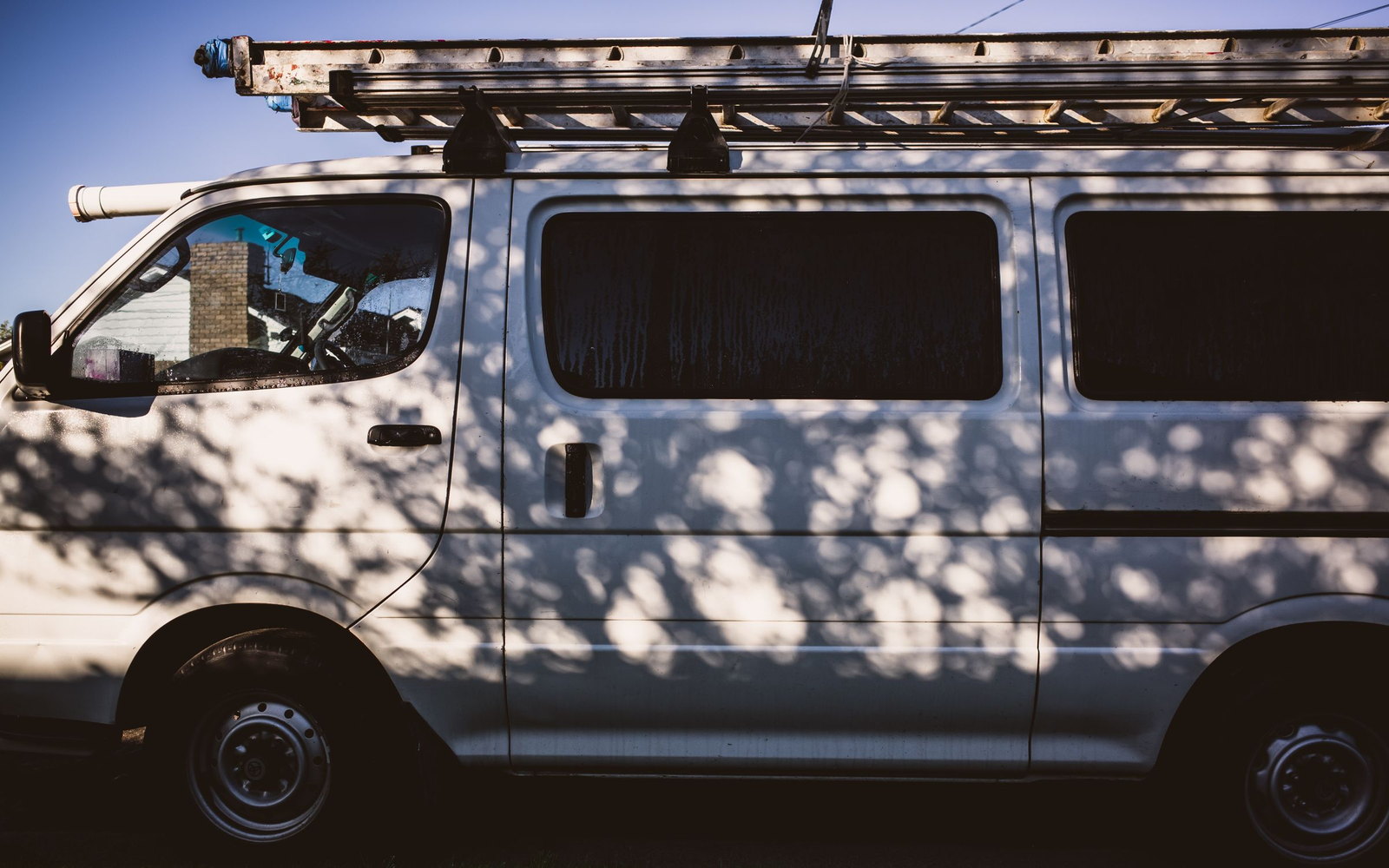 A plain white van with a ladder on the roof rack, tree shadow moving across its flank