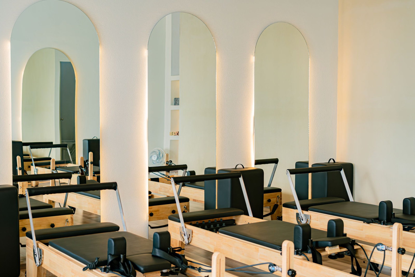 A row of pilates reformers in an empty studio, backlit arched mirrors on the wall behind