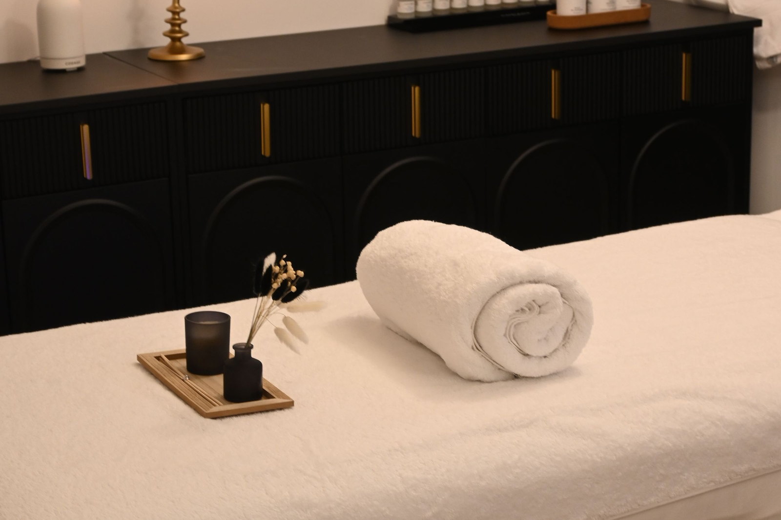 A treatment couch made up with a rolled towel and a tray of oils, a fluted black console behind